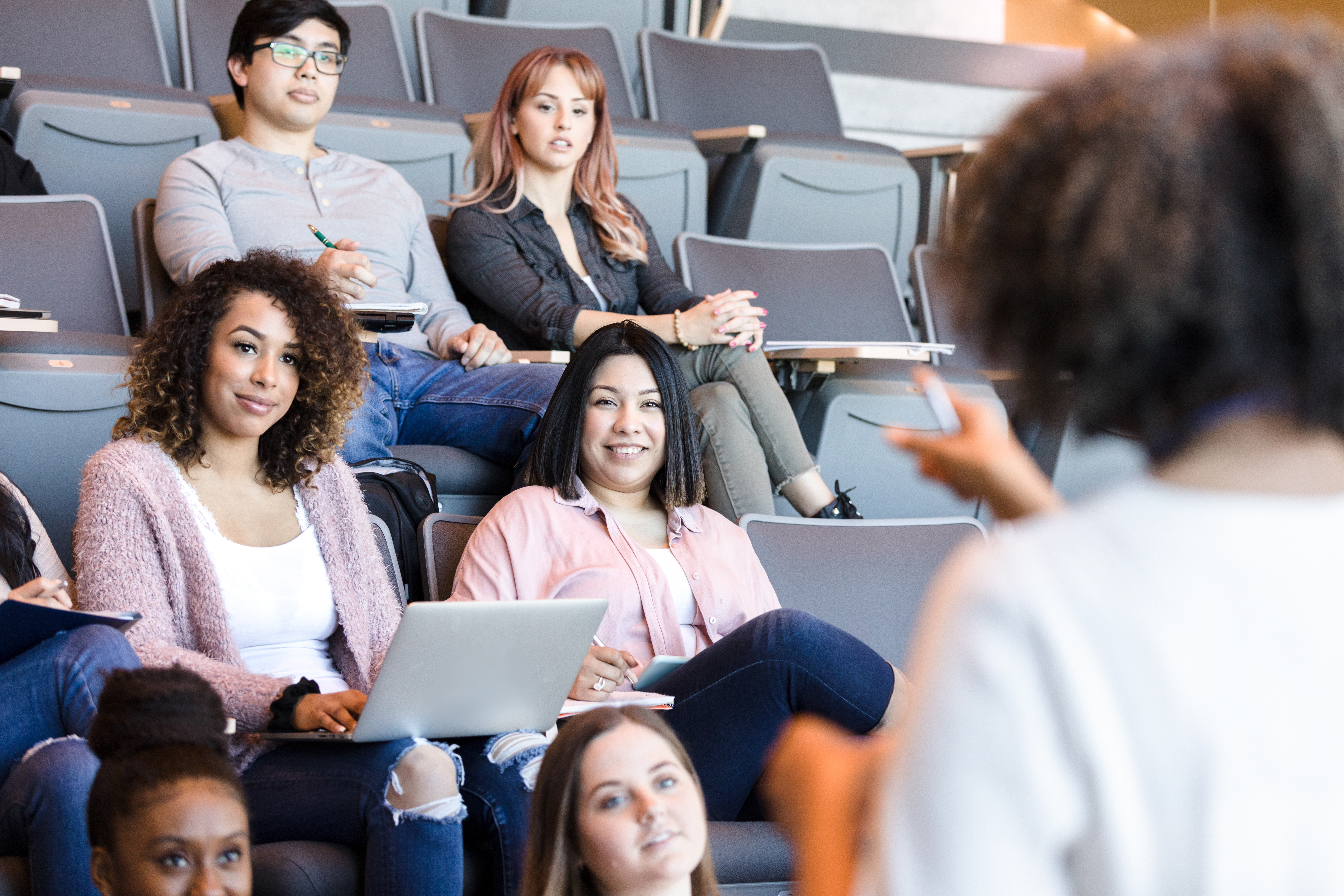 A female college professor gestures while giving a lecture. Focus is on the students listening attentively to the lecture.
