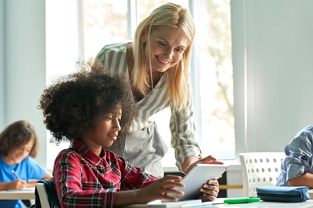 Happy mature female teacher educator helping African American junior school kid girl student using digital tablet computer education program app technology during elementary class lesson in classroom.; Shutterstock ID 2036186273; purchase_order:DPG Bulk Purchase Jul/Aug 2025; job:DPG selections; client:McGraw Hill; other: