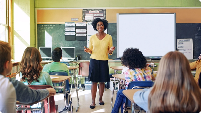 Standing at the front of a classroom, a teacher smiles as she speaks.