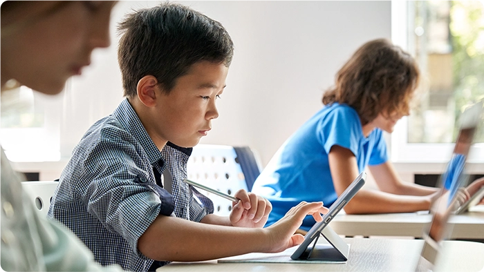 Three children sit at desks using tablet computers.